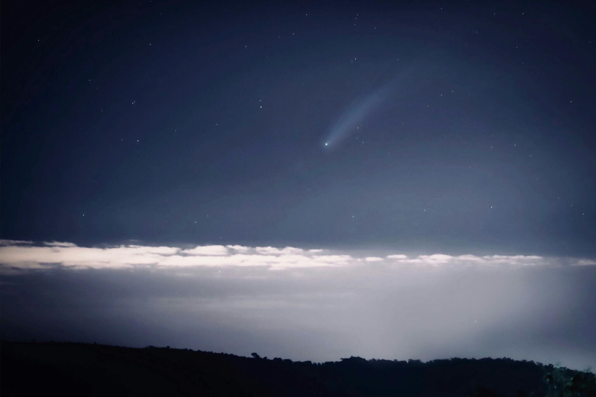 Astounding view of a luminous comet streaking across a starry night sky above a cloud layer.